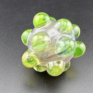 May include: A spherical glass bead featuring a clear, translucent core adorned with multiple raised, bright lime green circular bumps. The surface of the glass displays a smooth, polished finish with subtle internal light reflections throughout the structure.