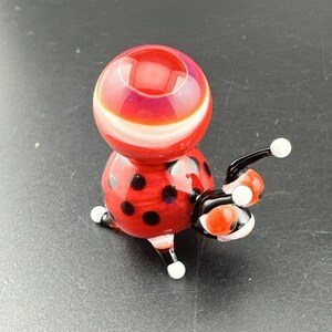 May include: A glass ladybug figurine featuring a bright red spherical body with black polka dots, a smooth red head section with a white horizontal band, and black legs and antennae tipped with small white spheres for a decorative accent.