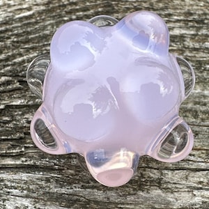 May include: A translucent, pale lavender glass knob with a rounded, multi-lobed flower-like shape. The surface is smooth and glossy, featuring a soft, milky opalescent quality that catches the light, creating subtle highlights across its curved form.