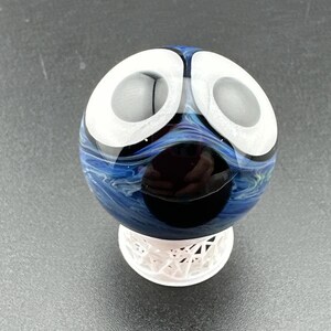 May include: A decorative glass orb with a unique design. The orb features a combination of white, blue, and black colors. It is supported by a white, geometric base. The glass has a swirling pattern, creating a visually appealing effect.