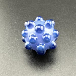 May include: A blue and white glass bead with a unique, textured surface. The bead is spherical, with numerous rounded protrusions. The color is a mix of light blue and white, creating a visually interesting pattern. The bead is likely handmade.