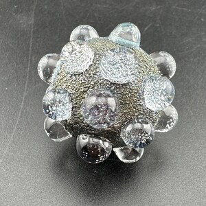 May include: A spherical glass object with a textured, dark metallic gray core. The surface is covered in multiple clear, raised glass bubbles of varying sizes, each containing tiny air inclusions that create a speckled, light-reflecting appearance.