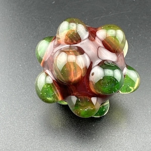 Pode incluir: Uma escultura de vidro esférica com um grupo de nós arredondados e salientes. A superfície apresenta uma mistura de tons verde translúcido profundo e castanho-âmbar quente, com um acabamento liso e polido que reflete a luz na sua forma.