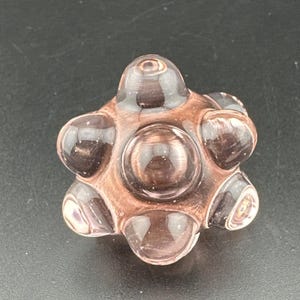 May include: A clear, spherical glass object with a starburst design. The object is a translucent, purplish-brown color with rounded, bubble-like protrusions. The central sphere is larger than the surrounding ones. The glass has a smooth, polished appearance.