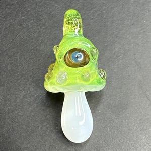 May include: A whimsical, hand-blown glass mushroom pendant. The mushroom cap is a vibrant green with textured details and a single blue eye. The stem is a smooth, opaque white. A unique, artistic piece.