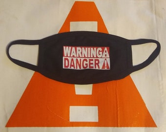Może przedstawiać: Czarna maska na twarz z napisem "WARNINGA DANGER!" w czerwonych i białych literach blokowych. Maska jest umieszczona na pomarańczowym i białym tle.