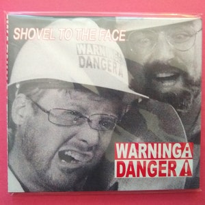 May include: A black and white photo of two men wearing glasses. The man in the foreground is wearing a white hard hat with the words "WARNINGA DANGER A" on it. The text "SHOVEL TO THE FACE" is at the top of the image.