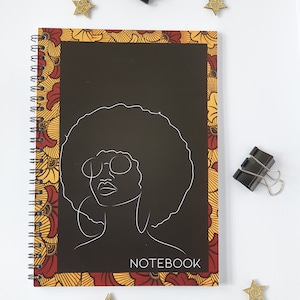 Può includere: Un quaderno nero con rilegatura a spirale e una copertina con un motivo colorato. La copertina presenta un disegno a linea di una donna con un afro e occhiali. Il testo "NOTEBOOK" è stampato sulla copertina.
