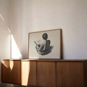Pu&ograve; includere: Un'illustrazione in bianco e nero di una persona seduta e che legge un libro. L'illustrazione &egrave; incorniciata e appesa a un muro sopra un armadio in legno.
