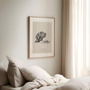 May include: A black and white illustration of a dog looking up at a plate with a piece of cake on it. The illustration is framed in a light wood frame and is hanging on a light beige wall.