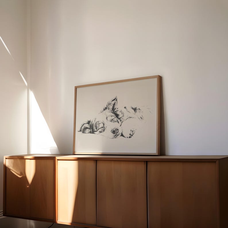 May include: A framed black and white sketch of a dog lying down. The artwork is in a light wood frame and rests on a wooden cabinet. The dog's features are detailed, with a focus on its face and paws.