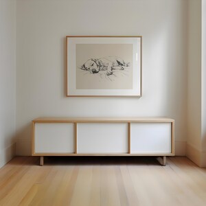 May include: A white cabinet with three sliding doors, made of wood with a light wood finish. The cabinet is sitting on a light wood floor. A framed black and white drawing of a dog lying down is hanging on the wall above the cabinet.