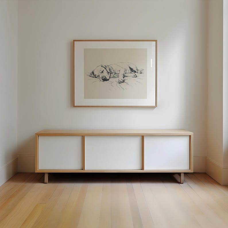 May include: A white cabinet with three sliding doors, made of wood with a light wood finish. The cabinet is sitting on a light wood floor. A framed black and white drawing of a dog lying down is hanging on the wall above the cabinet.