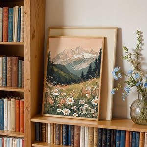 May include: A framed print of a mountain landscape with a field of wildflowers in the foreground. The mountains are snow-capped and the sky is a soft pink. The print is hanging on a wall with a bookshelf to the left and a vase of blue flowers to the right.