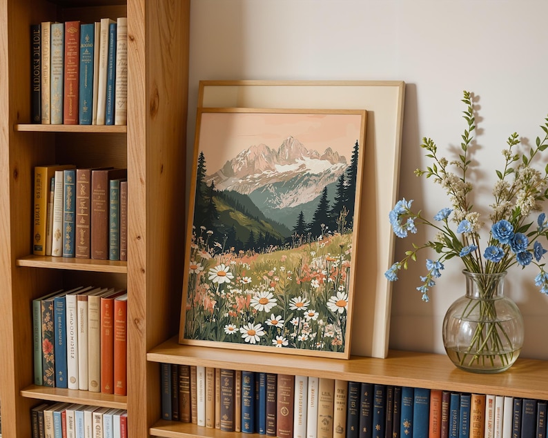 May include: A framed print of a mountain landscape with a field of wildflowers in the foreground. The mountains are snow-capped and the sky is a soft pink. The print is hanging on a wall with a bookshelf to the left and a vase of blue flowers to the right.