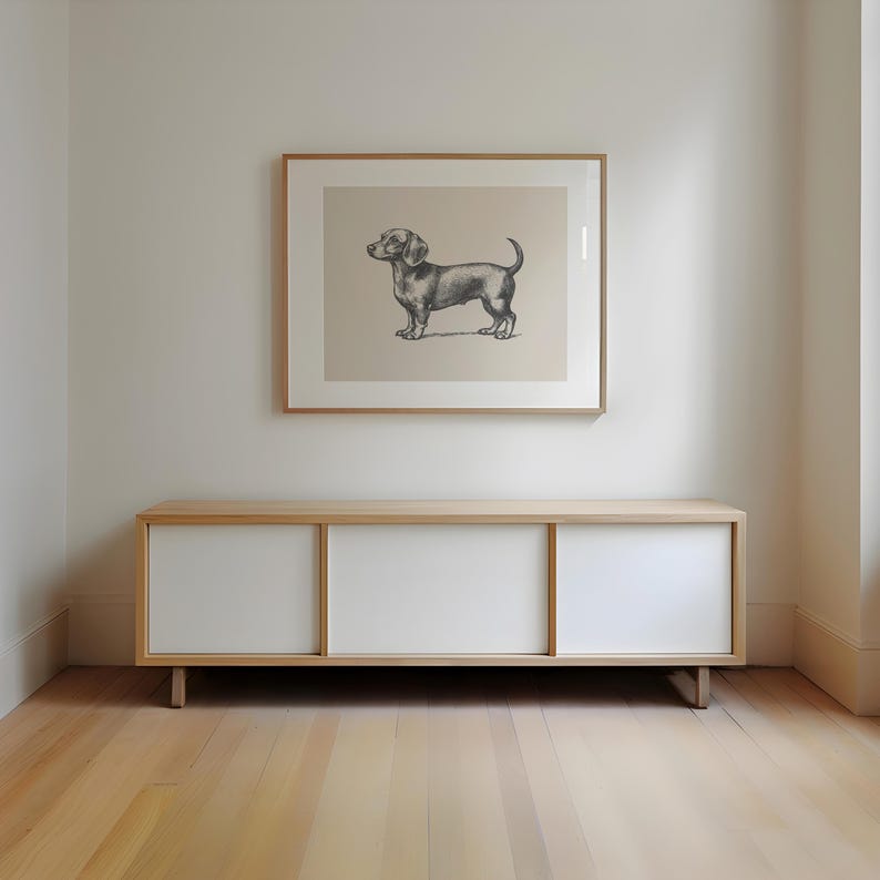 Peut inclure: Un cadre avec une illustration de chien teckel est suspendu au-dessus d'un meuble bas et moderne. Le meuble a un cadre en bois clair avec des portes coulissantes blanches. La pi&egrave;ce a un parquet clair et des murs blancs.