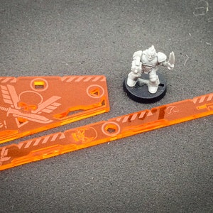 May include: Two orange acrylic gaming tokens with a skull and wings design. One token has the word "Epic!" on it. The tokens are on a gray surface with a miniature grey plastic figure.