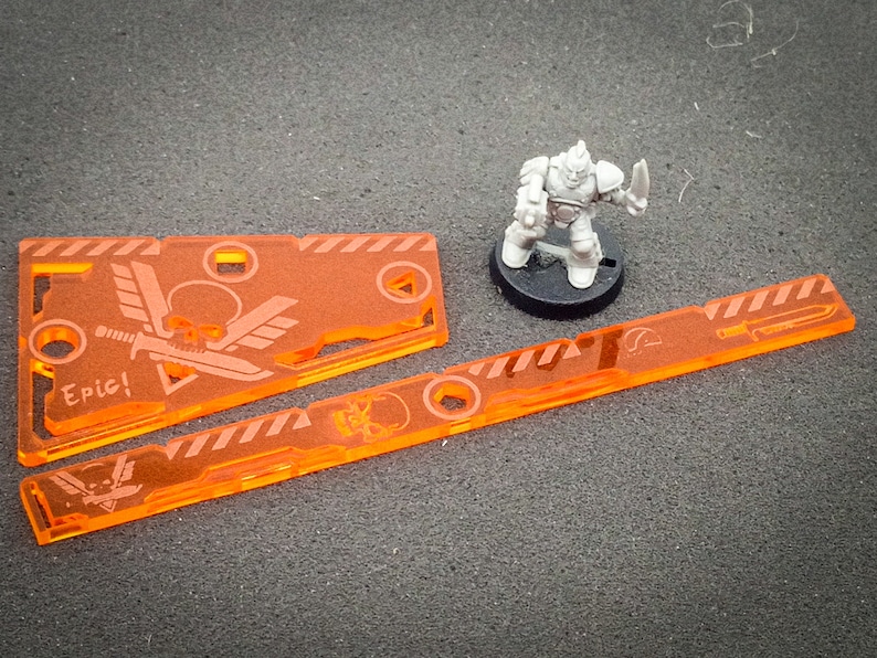 May include: Two orange acrylic gaming tokens with a skull and wings design. One token has the word "Epic!" on it. The tokens are on a gray surface with a miniature grey plastic figure.