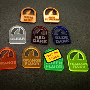 May include: Assortment of 10 shark fin shaped acrylic tokens in various colors, including clear, red, blue, orange, and yellow. Some tokens are fluorescent. The tokens are labeled with the material and thickness, including 3mm MDF, 4mm plywood, and various colors. One token is labeled "Out of Stock".