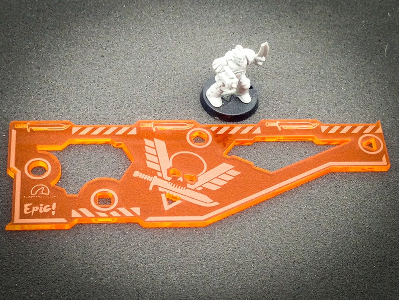 May include: Orange plastic gaming token with a skull and wings design. The token has the word "Epic!" printed on it. A miniature space marine figure is standing next to the token.