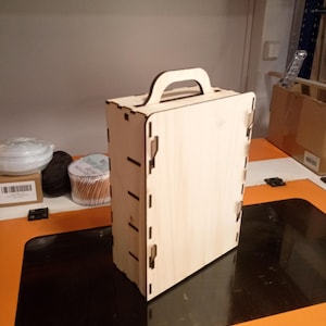 Può includere: Una scatola di legno color chiaro con manico, probabilmente per riporre o trasportare oggetti. La scatola è costruita con pezzi ad incastro e ha una forma rettangolare. Il manico è arcuato e realizzato nello stesso legno chiaro. La scatola è appoggiata su una superficie nera e arancione.