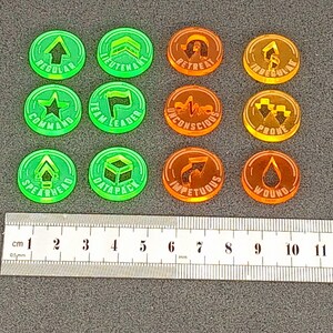 May include: Twelve round, plastic tokens with various symbols and text. The tokens are green and orange. The text on the tokens includes "Regular", "Lieutenant", "Retreat", "Irregular", "Command", "Team Leader", "Unconscious", "Prove", "Spearhead", "Datapack", "Impetuous", and "Wound".