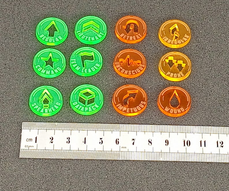 May include: Twelve round, plastic tokens with various symbols and text. The tokens are green and orange. The text on the tokens includes "Regular", "Lieutenant", "Retreat", "Irregular", "Command", "Team Leader", "Unconscious", "Prove", "Spearhead", "Datapack", "Impetuous", and "Wound".