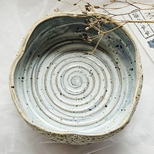 May include: A round ceramic bowl with a light blue and white glazed interior featuring a spiral pattern and dark speckles. The exterior has a textured, rough surface. A dried plant stem rests across the top, next to a small white tag with text.
