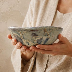 Może przedstawiać: Ręcznie wykonana ceramiczna miska o nieregularnym, organicznym brzegu. Zewnętrzna strona posiada nakrapiane szkliwo w odcieniach przygaszonego błękitu, szarości i kremu, z ciemnobrązowymi liniami przypominającymi spękania na powierzchni.