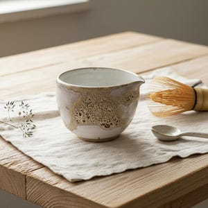Op de afbeelding: Een witte keramische matcha kom met een tuit en getextureerde bruine accenten. Een bamboe klopper en een kleine lepel liggen op een linnen doek. De kom staat op een houten tafel.