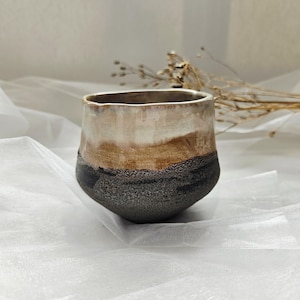 May include: A ceramic cup featuring a rounded shape and a multi-toned glaze. The upper portion is a smooth, creamy off-white, transitioning into a warm tan middle section, and finishing with a dark, textured, speckled charcoal base.
