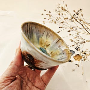 Op de afbeelding: Een kleine keramische kom met een uitlopende rand, voorzien van een reactief glazuur in tinten crème, beige en blauw. De buitenkant van de voet heeft een donkere, getextureerde bruine afwerking, terwijl de binnenkant verticale strepen en een blauw centrum toont.
