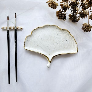 Op de afbeelding: Witte keramische schaal in de vorm van een ginkgo-blad met een gouden rand. De schaal staat op een wit oppervlak met drie kwasten.