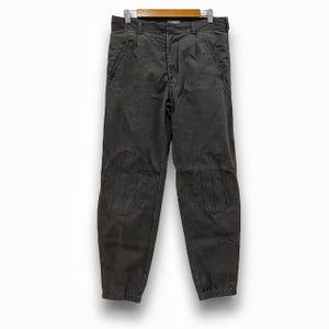 May include: Dark grey trousers with knee patches, hanging on a wooden hanger. The trousers have a button and zip closure, belt loops, and pockets. The bottom of the trousers have an elastic cuff.