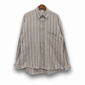 Puede incluir: Una camisa de manga larga con rayas verticales en tonos marrón, beige y azul. La camisa tiene un bolsillo en el pecho y está colgada de una percha de madera. El cuello está abotonado y los puños son visibles.