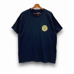 May include: A navy blue t-shirt with a small sunflower design on the front. The sunflower is yellow and green. The shirt is hanging on a wooden hanger.