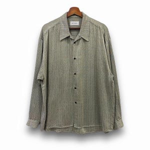 May include: A long-sleeved button-down shirt in a textured, light olive green fabric. The shirt features a classic collar, black buttons, and the brand name "SMASH" on a small label near the collar. The shirt is hanging on a wooden hanger.