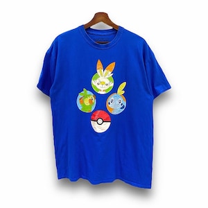 May include: A blue t-shirt with a graphic of four Pokemon characters from the Sword and Shield video game series. The characters are Grookey, Scorbunny, Sobble, and a Pokeball.