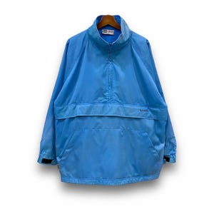 Puede incluir: Una chaqueta cortavientos azul claro con cierre de cremallera a la mitad y un bolsillo canguro. La chaqueta tiene un pequeño logotipo en el pecho izquierdo y un logotipo más grande en el lado derecho del bolsillo.
