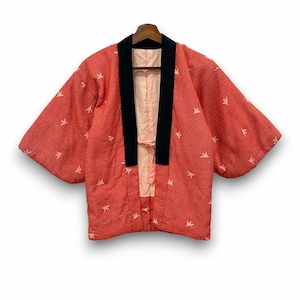May include: A quilted jacket in a vibrant coral-red hue featuring a repeating geometric pattern and small white bird motifs. The garment has wide sleeves, a solid black collar, and a light pink interior lining with a thin fabric tie closure at the front.