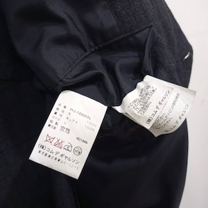 Op de afbeelding: Een wit label met zwarte tekst die de samenstelling van een kledingstuk beschrijft. Het label luidt "HJ-10005L", "100% wol", "100% cupro", "Maat L", "AD1999", "(株)コムデギャルソン", "東京都港区南青山5-11-5".