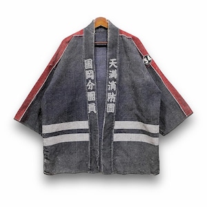 Puede incluir: Una chaqueta de bombero japonesa azul oscuro con detalles rojos en las mangas. La chaqueta presenta rayas horizontales blancas y texto japonés. Un parche circular es visible en la manga derecha. La chaqueta está colgada en una percha de madera.