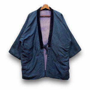 Puede incluir: Chaqueta azul estilo kimono con cuello y ribete de terciopelo negro, colgada de una percha de madera. La chaqueta tiene un tejido texturizado y un forro morado. Cuenta con mangas anchas y cierre de lazo en la parte delantera.