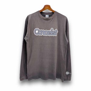Pode incluir: Uma camiseta cinza de manga comprida com a palavra "Canoeist" impressa em branco e cinza na frente. A camiseta tem gola redonda e um caimento solto.