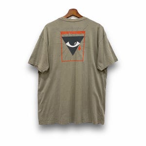 May include: A khaki-colored t-shirt with a graphic on the back. The graphic features a black triangle with an eye, a red square border, and the words "KNOW TOMORROW" in orange. The shirt is hanging on a wooden hanger.