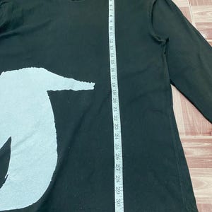 Puede incluir: Camiseta negra de manga larga con un gran gr&aacute;fico blanco en la parte delantera. La camisa se mide con una cinta m&eacute;trica, mostrando una longitud de aproximadamente 84 cm. Tiene cuello redondo y mangas largas.