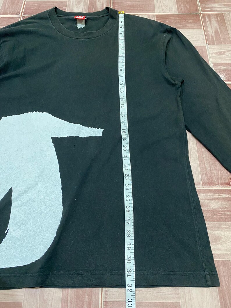 Puede incluir: Camiseta negra de manga larga con un gran gr&aacute;fico blanco en la parte delantera. La camisa se mide con una cinta m&eacute;trica, mostrando una longitud de aproximadamente 84 cm. Tiene cuello redondo y mangas largas.