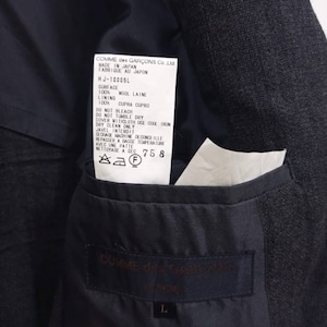 Op de afbeelding: Een zwarte blazer met een label met de tekst "COMME des GAR&Ccedil;ONS Co., Ltd. MADE IN JAPAN FABRIQUE AU JAPON HJ-10005L SURFACE 100% WOOL LAINE LINING 100% CUPRA CUPRO DO NOT BLEACH DO NOT TUMBLE DRY COVER WITH CLOTH USE COOL IRON DRY CLEAN ONLY JAVEL INTERDIT SECHAGE MACHINE DE CONSEILLE REPASSER A BASSE TEMPERATURE AVEC UNE PATTE NETTOYAGE A SEC 758 F".