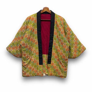 May include: A vintage kimono-style jacket with a geometric pattern in shades of green, orange, and yellow. The jacket has a black collar and trim, and a red interior. It is hanging on a wooden hanger.