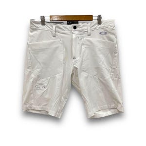 May include: White athletic shorts with multiple pockets and a button closure. The shorts are hanging on a wooden hanger. The Oakley logo is visible on the front. The shorts have a modern, sporty design.